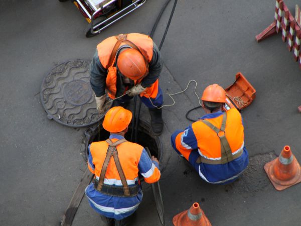 Drei Arbeiter mit Schutzhelmen an geöffnetem Kanaldeckel
