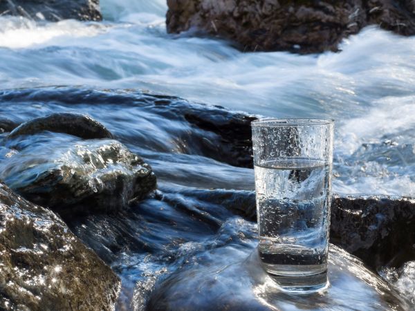Klares Wasser in einem Glas, das auf einem mit Wasser überspülten Stein steht. Er befindet sich in einem Bachlauf mit vielen Steinen.