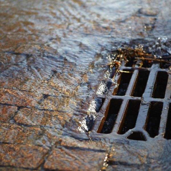 Abwasser läuft in einen vergitterterten Einlaufschacht für Strassenabwässer am Rand einer gepflasterten Straße.