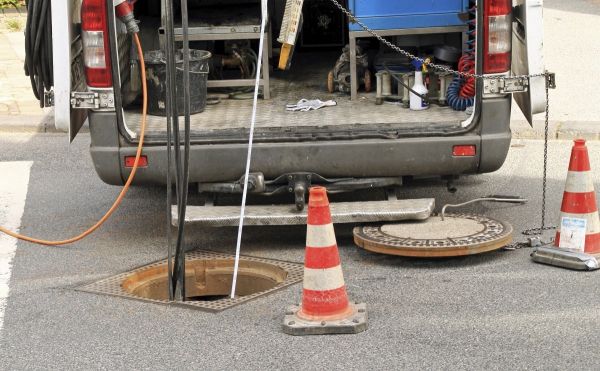 Ein Montagetransporter steht mit geöffneter Hecktüre und elektrisch angeschlossenen Werkzeugen direkt an einem geöffneten Kanaleinstieg/Gulli, in den Kabel führen. Die Baustelle ist mit 2 Pylonen abgesichert.