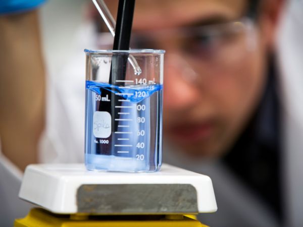 Ein Laborant mit Schutzbrille und Schutzhandschuhen tropft mit einer Pipette eine klare Füssigkeit in ein Messbecherglas mit anderer klarer Flüssigkeit, leicht blau gefärbt. Er rührt sie dabei gleichzeitig mit einem schwarzen Rührstab um. Das Becherglas s
